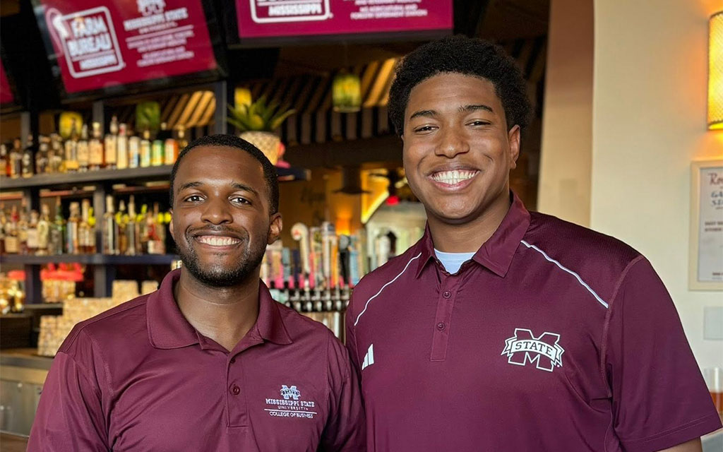 Two men wearing maroon Mississippi State University shirts smile while standing inside a bar or restaurant with bottles and TVs in the background.