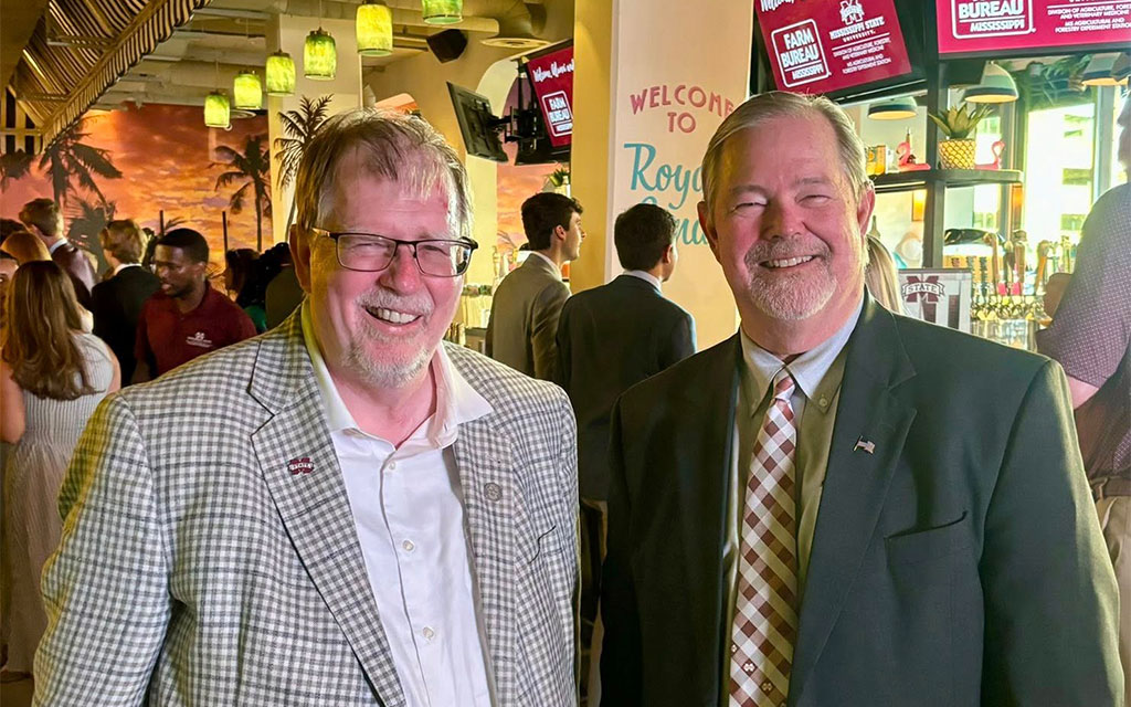 Two men in suits stand and smile at a social event inside a bar or restaurant, with people and a “Welcome to Royal” sign visible in the background.
