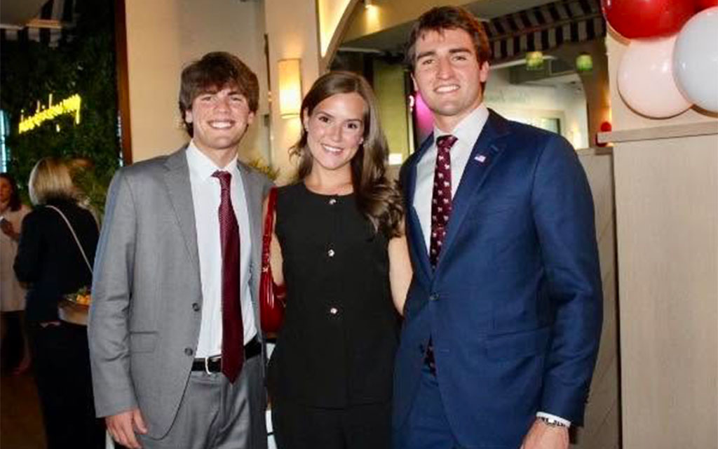Three young adults, two men in suits and a woman in a black outfit, pose together and smile at an indoor event decorated with white and red balloons.
