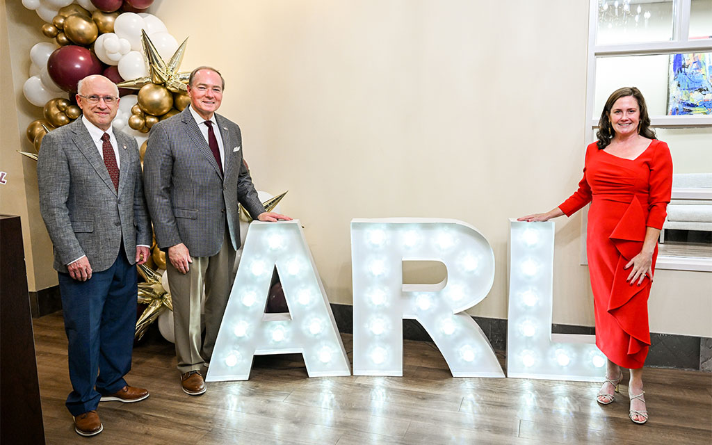 Three adults stand indoors beside large illuminated letters spelling “ARL,” with festive balloons in the background.