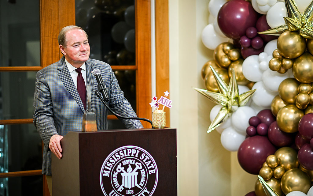 A man stands at a podium with the Mississippi State University seal, speaking into a microphone; festive balloons decorate the background.