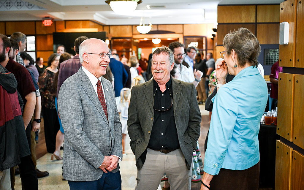 Three adults stand and talk in a busy indoor event space, smiling and dressed in business casual attire, with other people mingling in the background.