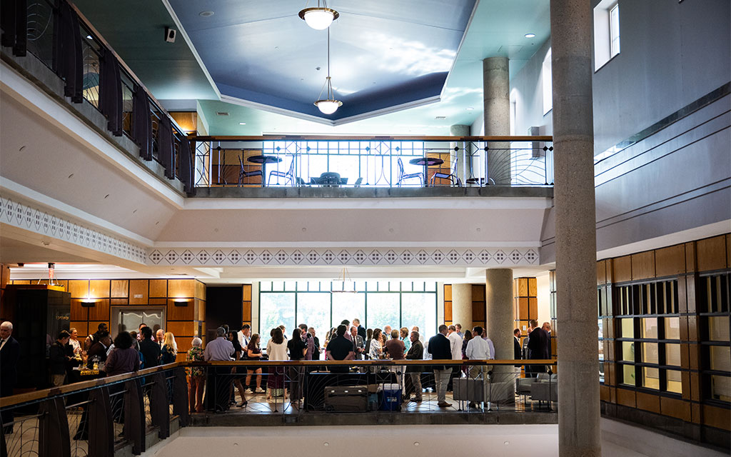 People gather and talk in a spacious two-level indoor atrium with large windows, overhead lighting, and modern architectural features.