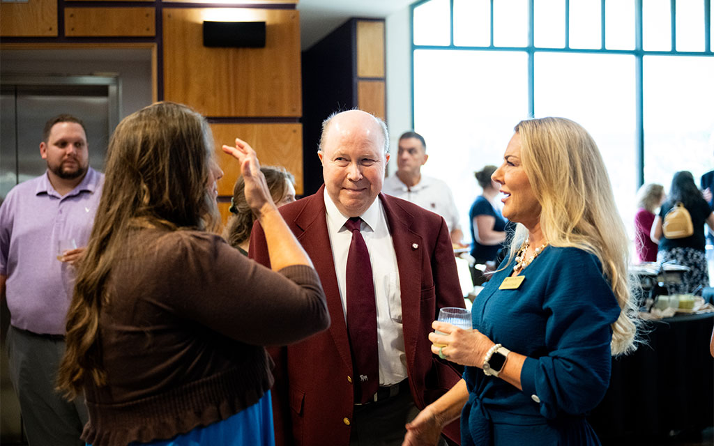 A group of people are talking at an indoor event. One man in a maroon jacket stands in the middle, while two women talk to him. Others are mingling in the background.