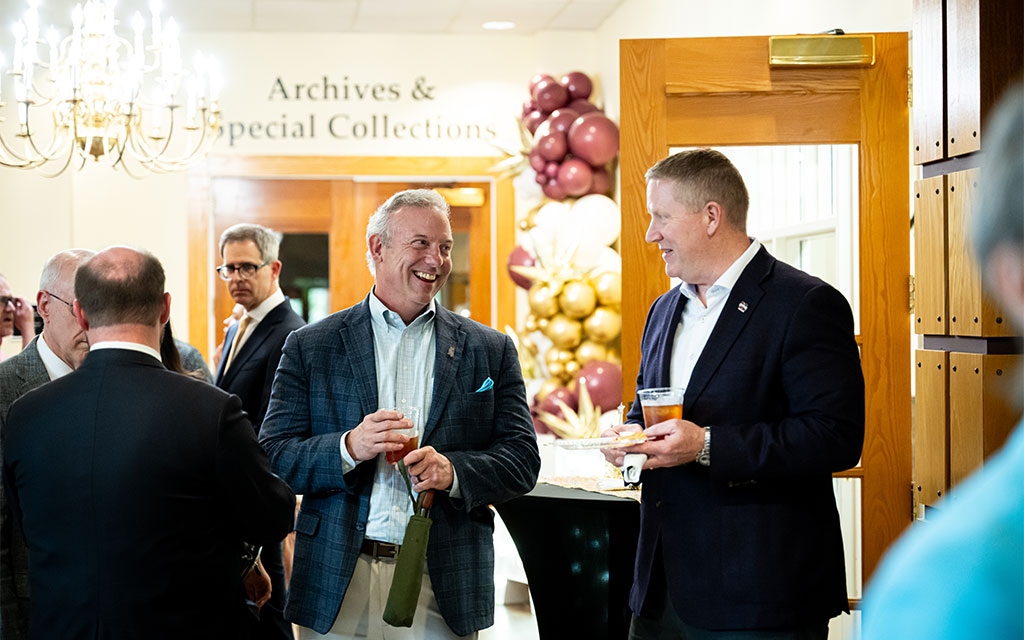 Several men in suits converse and smile at an indoor event near a sign reading “Archives & Special Collections,” with balloon decorations in the background.
