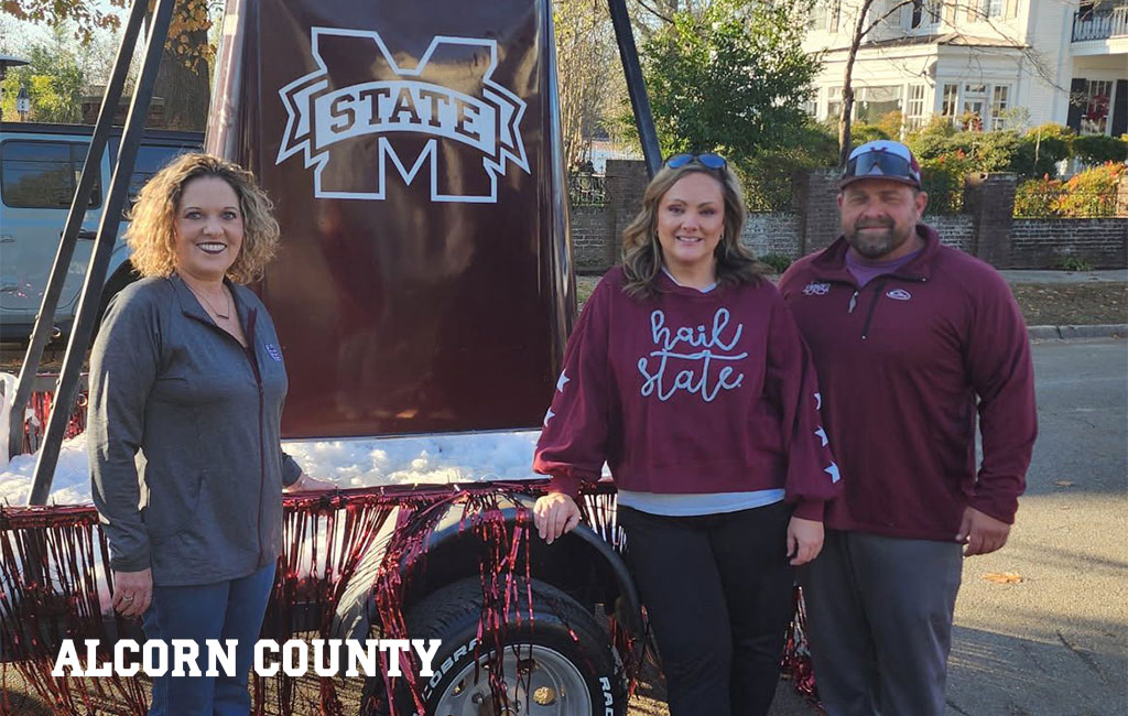 Three people stand in front of a maroon MS State display at an outdoor event. One wears a “hail state” shirt. The words “ALCORN COUNTY” appear in white text at the bottom left.
