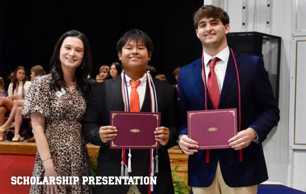 Three students stand indoors, two holding certificates, dressed in formal attire with medals and cords, at a scholarship presentation event.