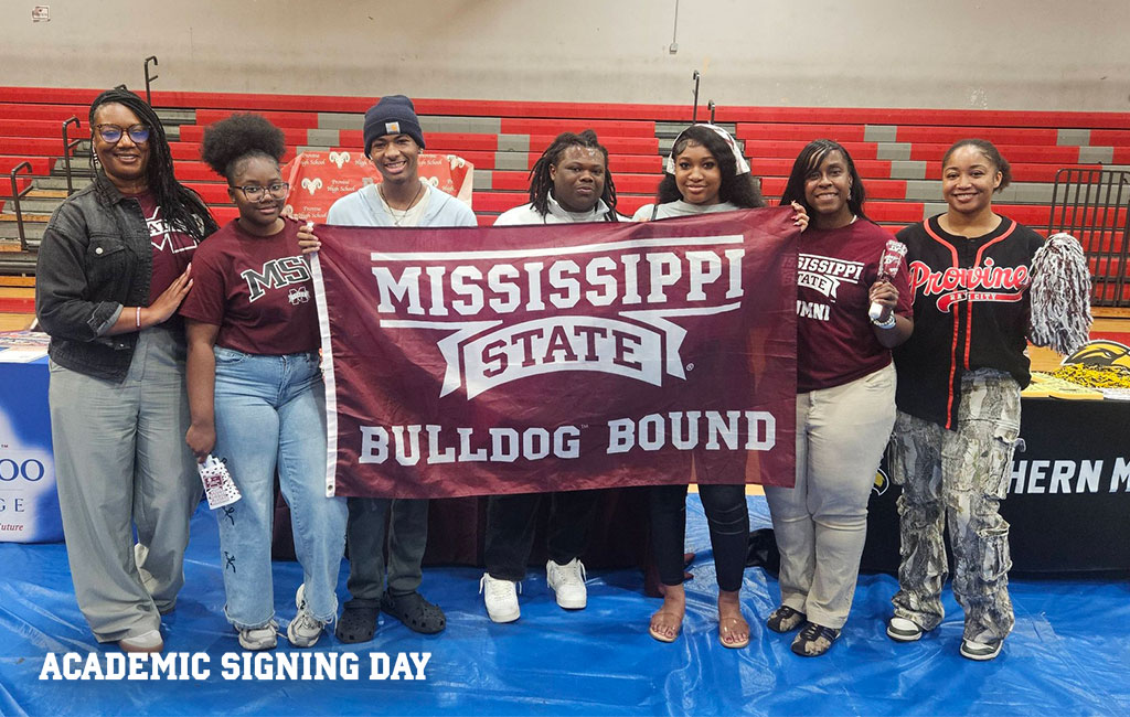 Seven people stand in a gym, holding a “Mississippi State Bulldog Bound” banner. They are smiling, wearing Mississippi State gear, and standing in front of tables on Academic Signing Day.