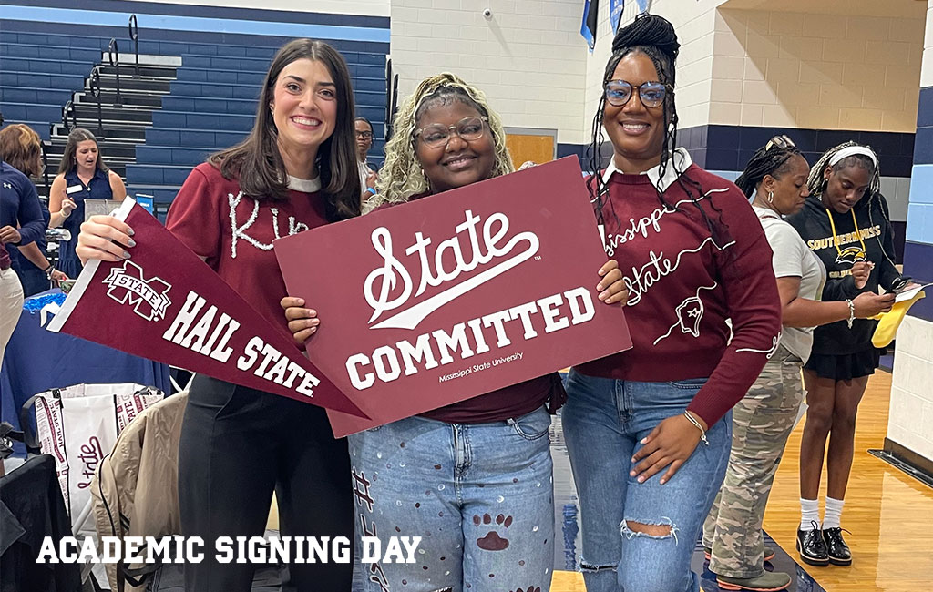 Three women pose indoors holding “Hail State” and “Committed” signs. Text reads “Academic Signing Day.” Other people are visible in the background.
