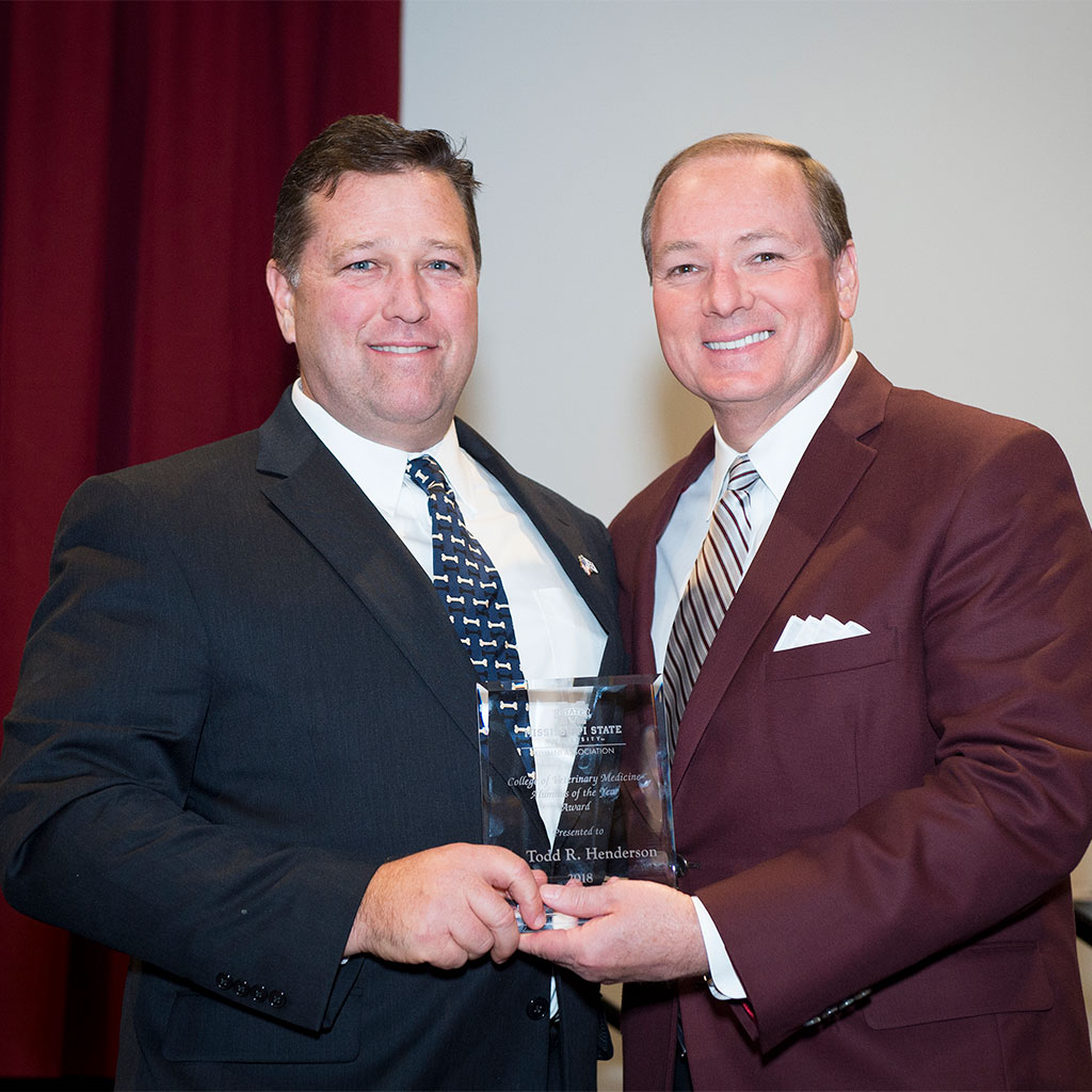Two men in suits stand together, smiling at the camera. One man is holding a clear award plaque. A red curtain and white wall are in the background.