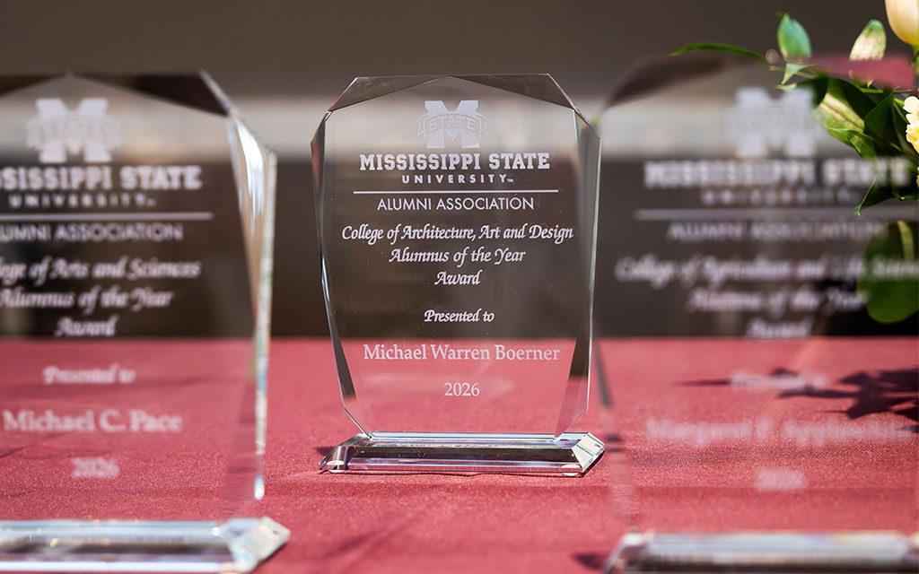 A clear glass award on a red tablecloth reads “Mississippi State University Alumni Association, presented to Michael Warren Boerner, 2026.” Other similar awards are visible in the background.