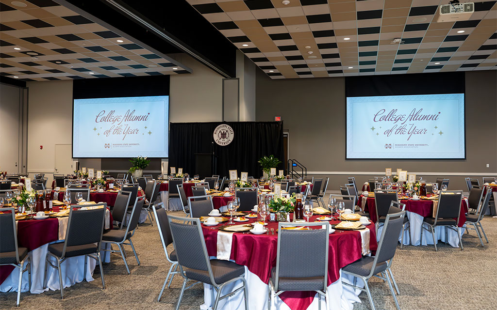 Banquet hall set up for an event, with round tables covered in red and white cloths, place settings, and two large screens displaying “College Alumni of the Year.”.