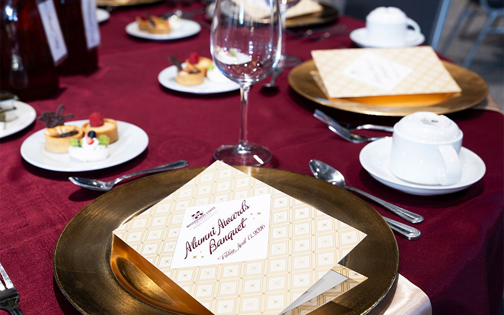 A banquet table set with gold plates, menus labeled “Alumni Awards Banquet,” wine glasses, desserts, and cups on a burgundy tablecloth.