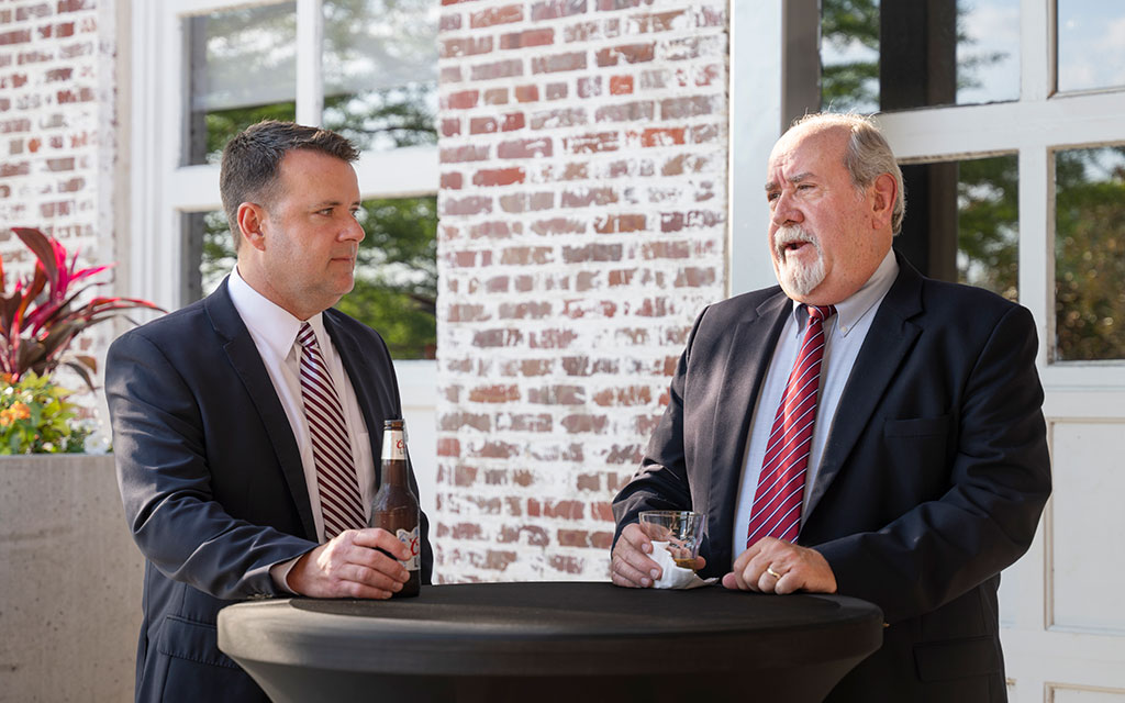 Two men in suits stand at a round table outdoors, one holding a bottle and the other a glass, engaged in conversation in front of a brick building.
