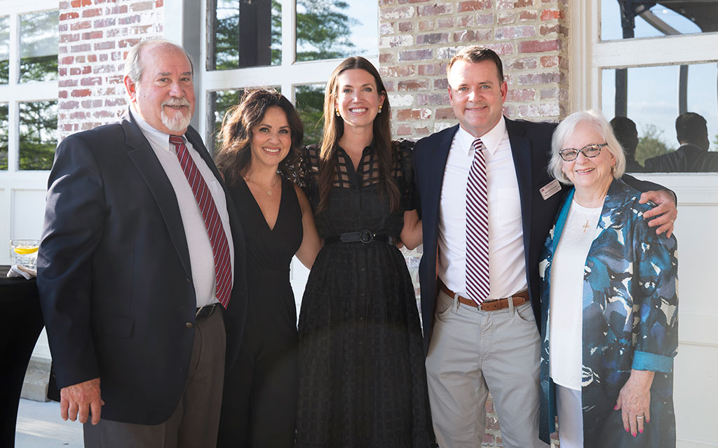 Five adults stand together, smiling, dressed in semi-formal attire, posing outdoors in front of a brick building with large windows.