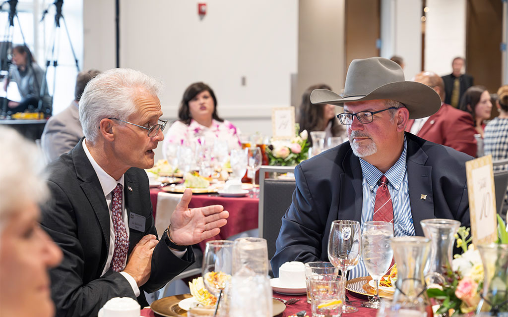 Two men in suits, one wearing a cowboy hat, sit at a round table with food and glasses, engaged in conversation at a formal event.