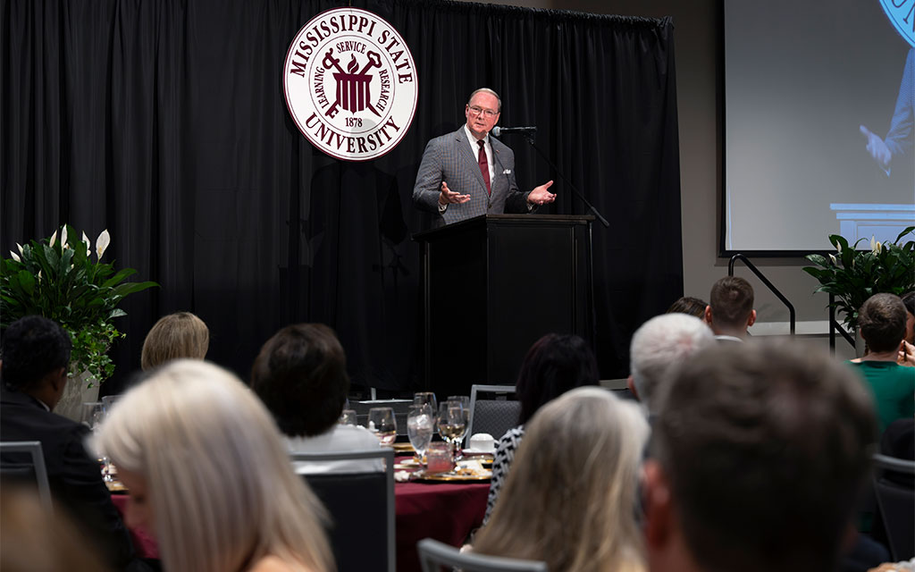A man in a suit speaks at a podium with the Mississippi State University logo behind him to an audience seated at dining tables.