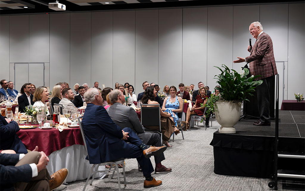 A man speaks on stage to a seated audience at a formal indoor event, with tables set for dining and attendees listening attentively.