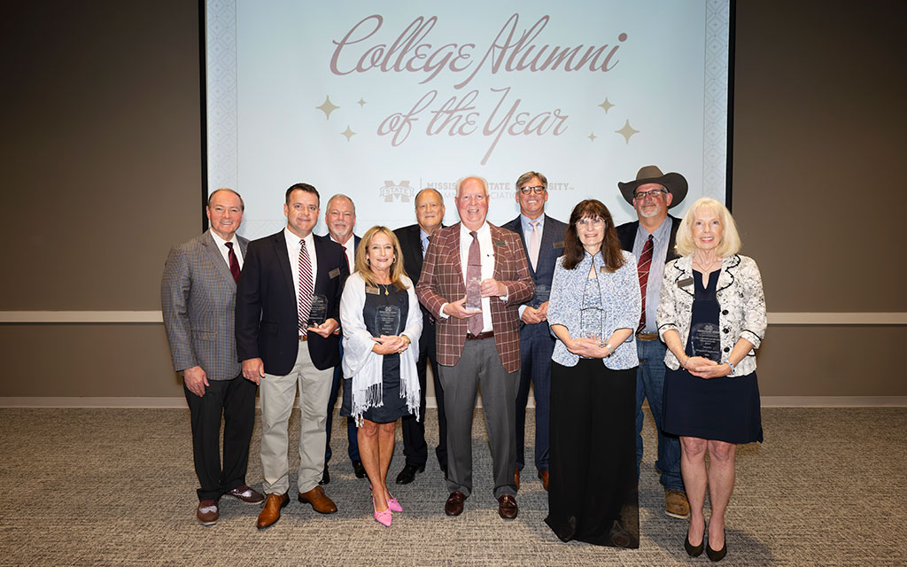 Ten people stand in a row holding awards in front of a screen that reads “College Alumni of the Year.”.