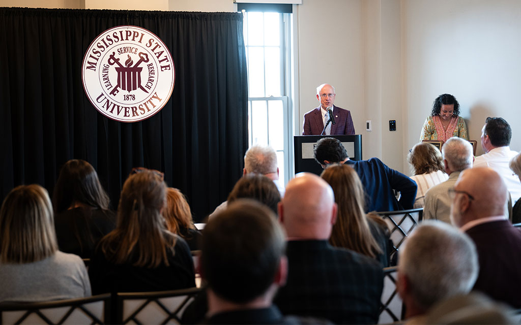 A speaker at a podium addresses an audience in a room with a Mississippi State University sign on a curtain in the background.
