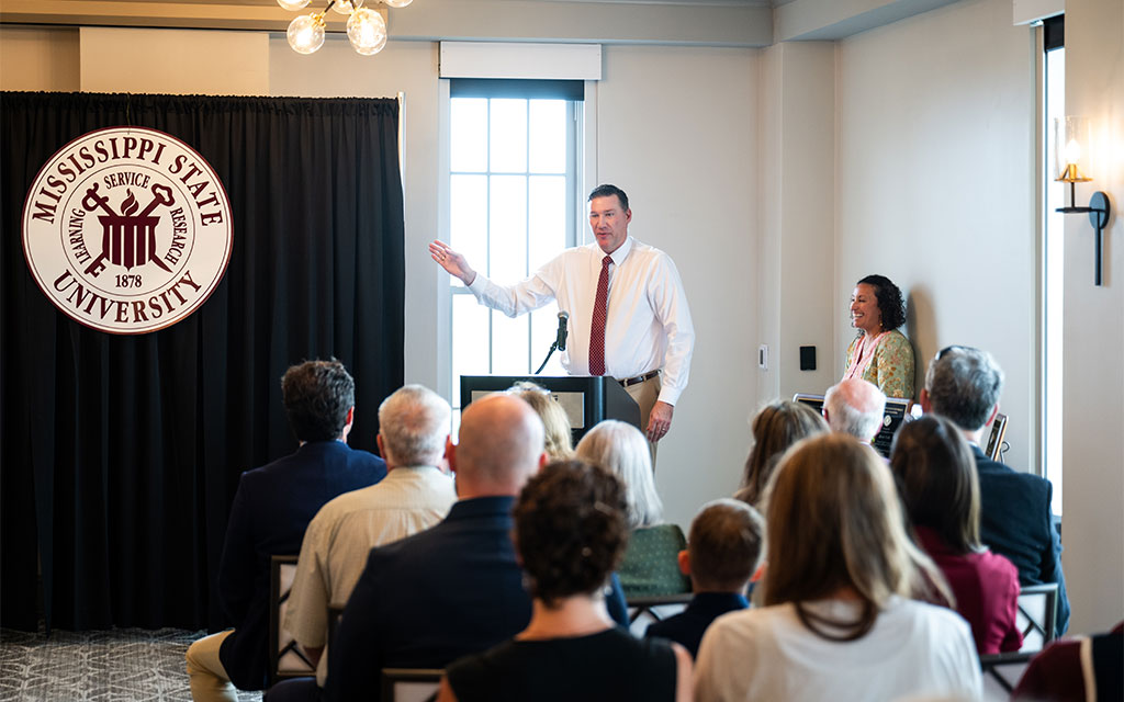 A man speaks at a podium to an audience in a room with a Mississippi State University banner. A woman stands nearby, smiling.