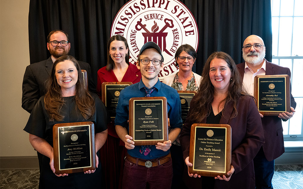 Seven people pose indoors holding plaques in front of a Mississippi State University emblem, suggesting an award or recognition ceremony.