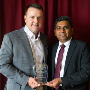 Two men in suits stand in front of a red curtain, holding a glass award together and smiling at the camera.