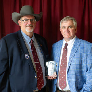 Two men in suits, one wearing a cowboy hat and holding an award, stand side by side in front of a red curtain.