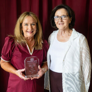 Two women stand in front of a maroon curtain; one holds a clear award plaque that reads “Mississippi State University.”.