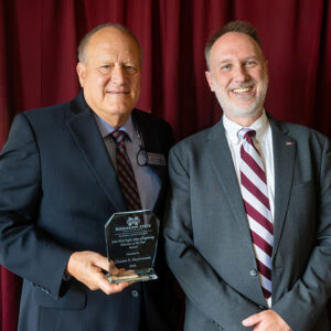 Two men in suits stand smiling; one holds an award plaque. A red curtain serves as the background.