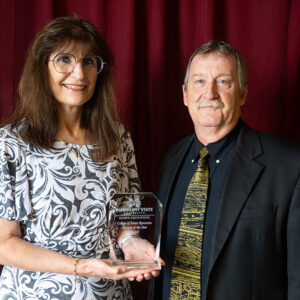 Two people stand in front of burgundy curtains; the woman holds an award plaque while both face the camera and smile.