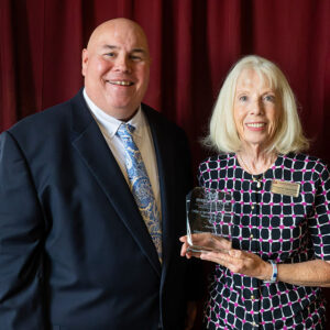 A man in a suit stands beside a woman holding a clear award plaque, both smiling, in front of a red curtain.