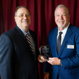 Two men in suits stand in front of a red curtain. One man holds a glass award. Both are smiling and facing the camera.