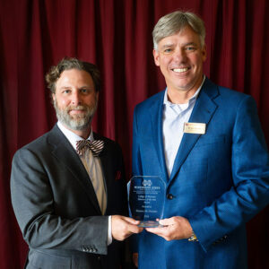Two men in suits stand in front of a red curtain; one holds a clear award plaque while both face the camera and smile.