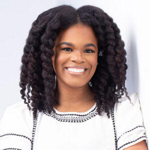 Smiling woman with natural, curly hair wearing a white shirt with black trim, standing against a light background.