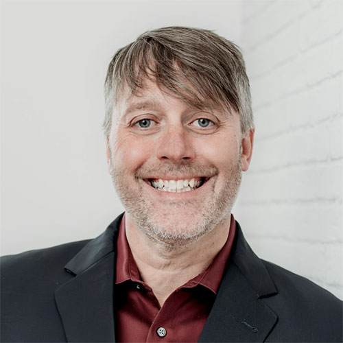 Smiling man with short, light brown hair and light stubble, wearing a dark blazer over a maroon shirt, standing in front of a white brick wall.