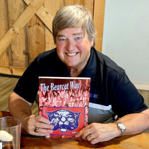 Smiling man with gray hair sits at a wooden table holding a book titled “The Bearcat Way,” which features a wildcat illustration and a crowd photo on the cover.