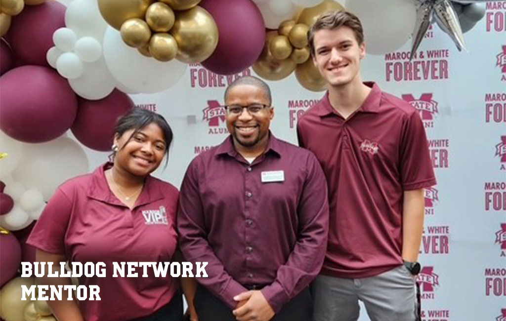 Three people wearing maroon shirts stand smiling in front of a balloon arch and a backdrop that says “MAROON & WHITE FOREVER” and “ALUMNI.” Text reads “BULLDOG NETWORK MENTOR.”.