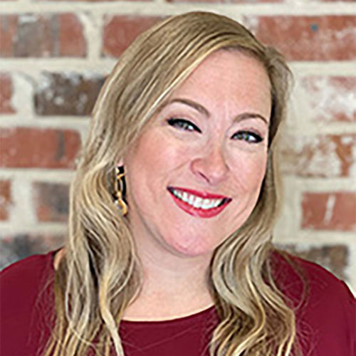 Smiling woman with long blonde hair wearing a red top stands in front of a brick wall background.
