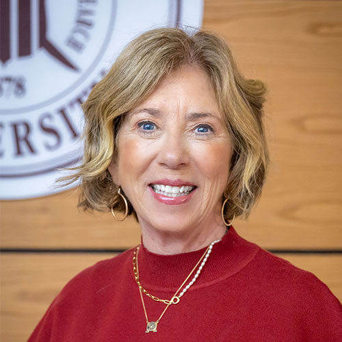 A woman with short blonde hair, wearing a red sweater and gold jewelry, smiles at the camera. A university seal is visible in the background on a wooden wall.