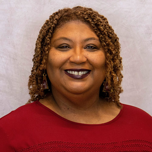 A woman with curly hair wearing a burgundy top and earrings smiles in front of a light gray background.