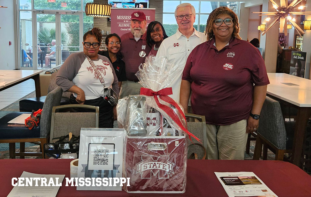 Six people stand behind a table with Mississippi State items, including a gift basket, flyers, and a QR code, at a Central Mississippi event in an indoor venue.