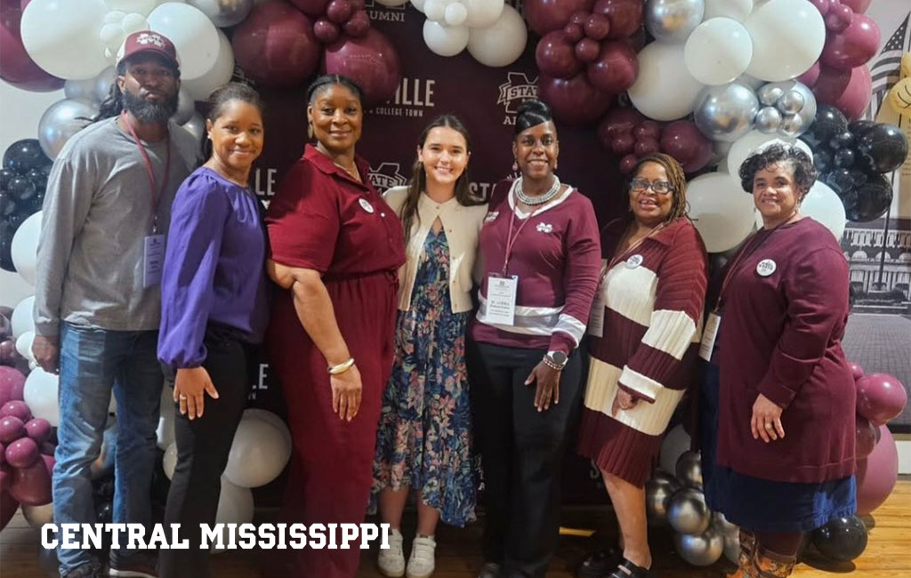 Seven people stand in front of a maroon and white balloon arch with a Central Mississippi sign and college-themed backdrop.