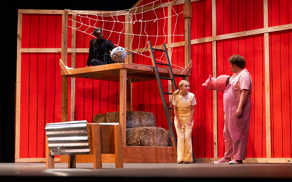 Three actors perform on stage with a red barn set; one in black sits in a web-like structure, another in yellow overalls looks up, and a third in pink holds a pig mask.