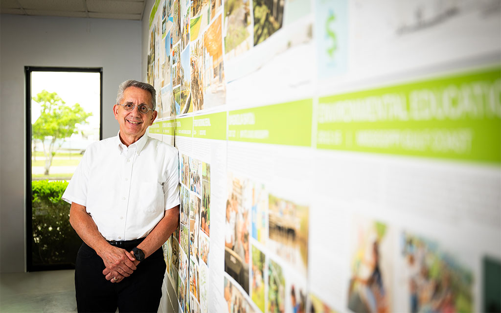 An older man in a white shirt stands smiling by a brightly lit wall covered with photos and text in an office space.