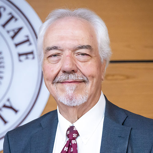 An older man with white hair and a mustache, wearing a suit and patterned tie, stands in front of a partial seal and wooden wall background.
