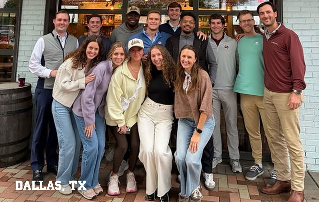 A group of thirteen adults, casually dressed, pose together and smile in front of a restaurant with a sign reading “Dallas, TX” in the lower left corner.
