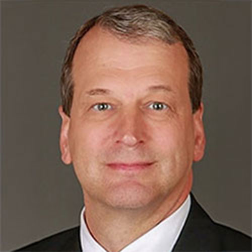 Middle-aged man with short gray hair, wearing a dark suit jacket, white shirt, and tie, posing against a plain gray background.
