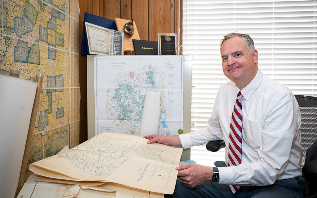 A man in a white shirt and striped tie sits at a desk examining large maps, with framed documents and maps displayed on the wall and behind him.