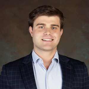 A man with short brown hair wearing a blue checked blazer and a light blue shirt smiles at the camera against a brown backdrop.